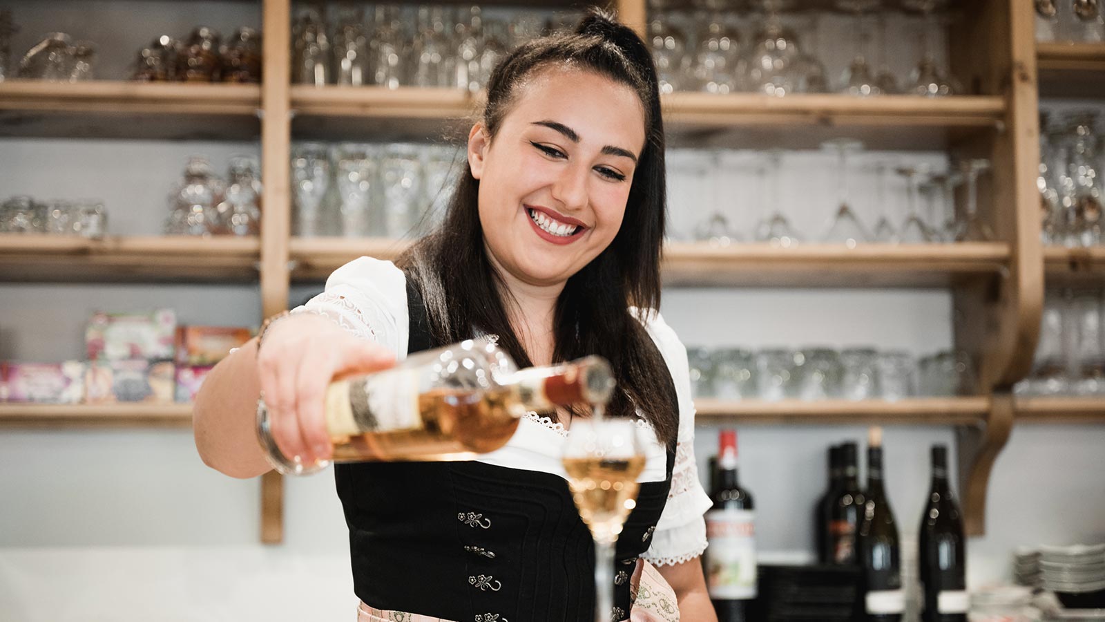 Una barista in vestito tradizionale versa un bicchierino di grappa al bancone del bar